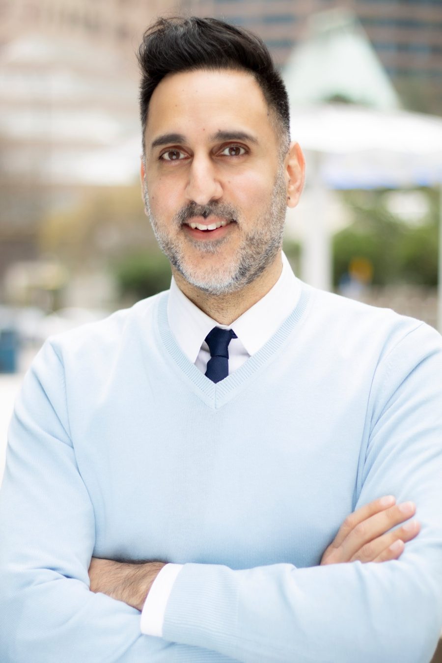 Portrait of Faizal Karim in a light blue sweater and navy tie, arms crossed, outdoors in downtown Vancouver